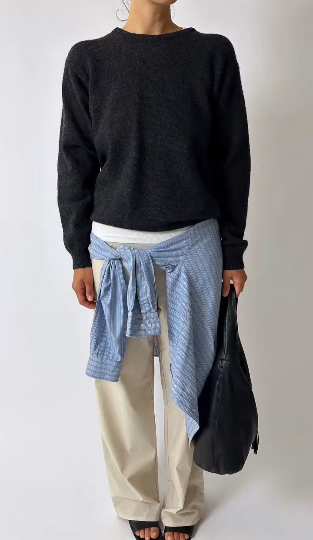 Person wearing a black sweater, light blue striped shirt, beige pants, and carrying a black bag on a white background