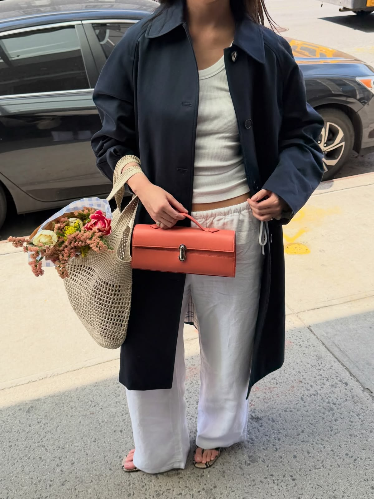 A woman standing outdoors on a sidewalk next to a street, wearing a long navy coat over a white tank top and white wide-leg pants, holding a coral-orange structured handbag and a woven tote bag filled with flowers.