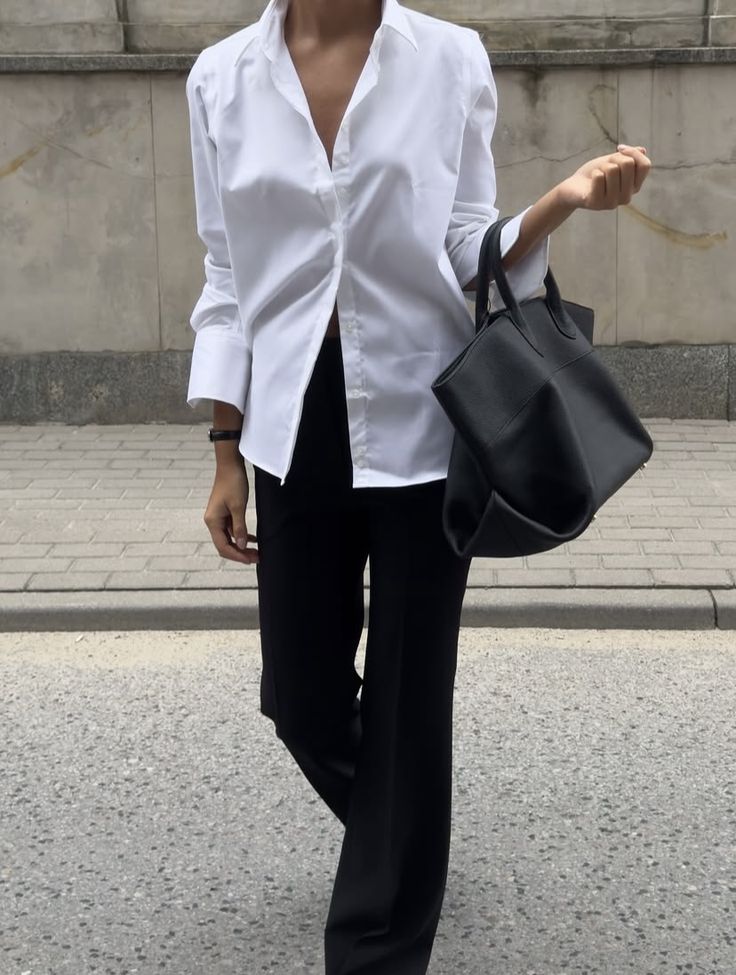 Person wearing a white shirt and black pants holding a black handbag on a street.