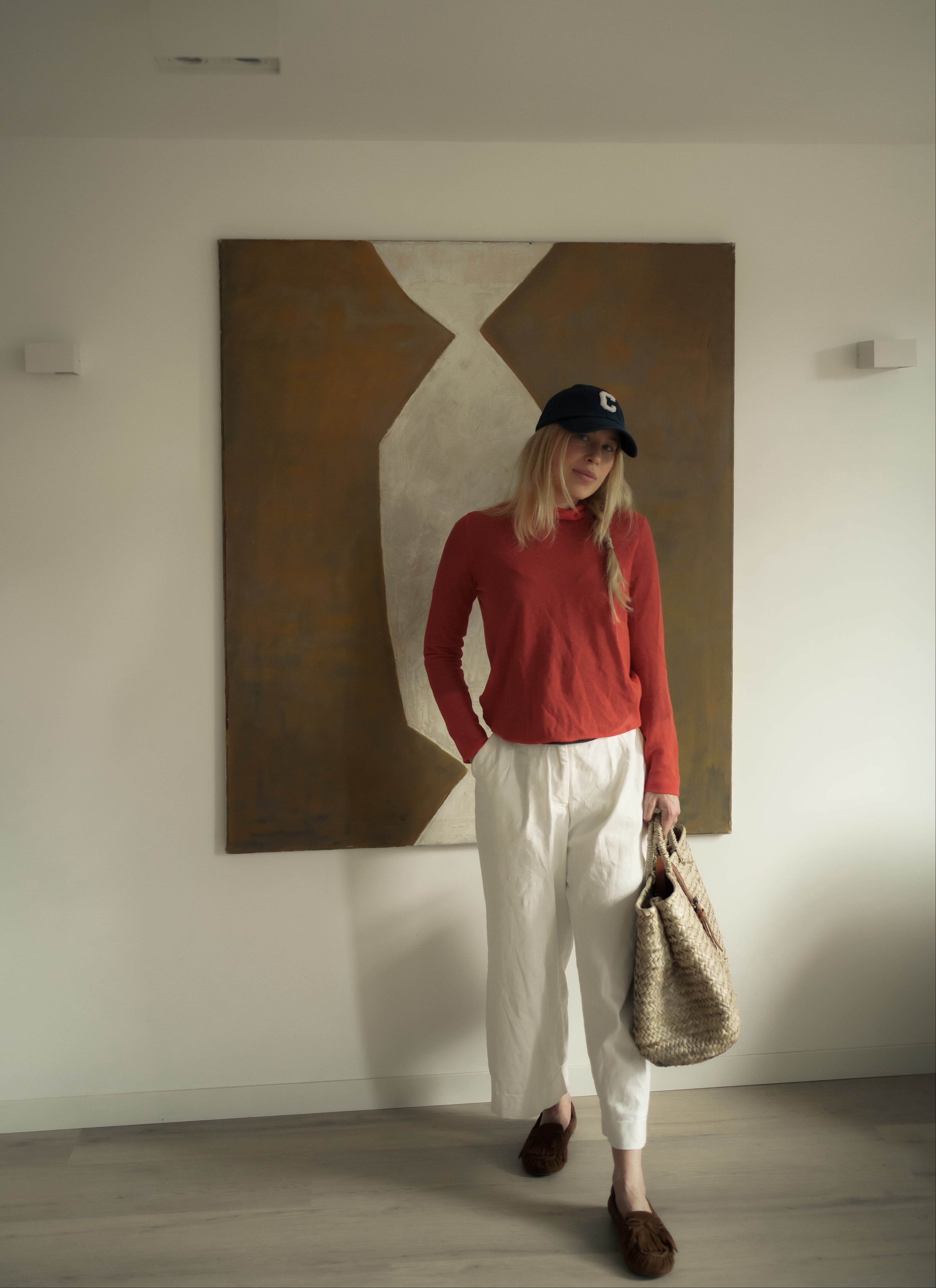 Woman in a red sweater and white pants standing in a room with a large abstract painting on the wall.