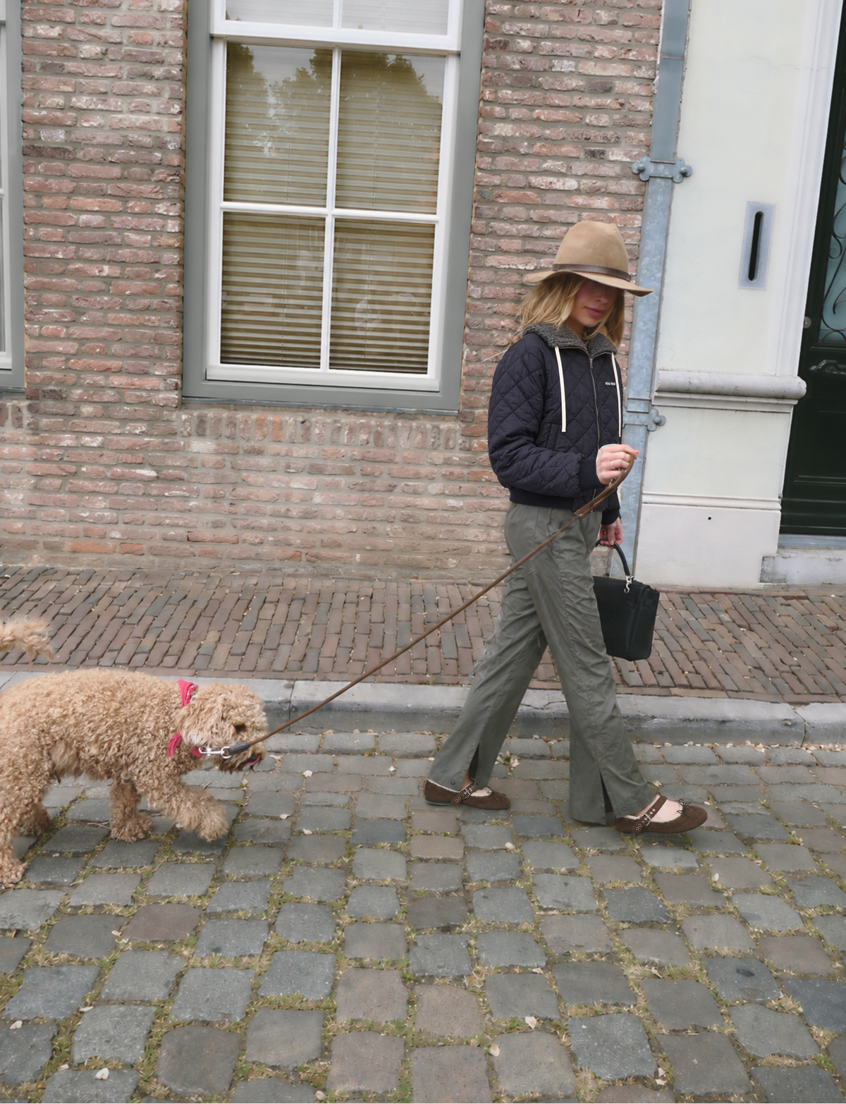 A woman wearing a wide-brimmed tan hat, dark navy jacket, olive green pants, and brown sandals walks a small beige poodle on a cobblestone street in front of a brick building.