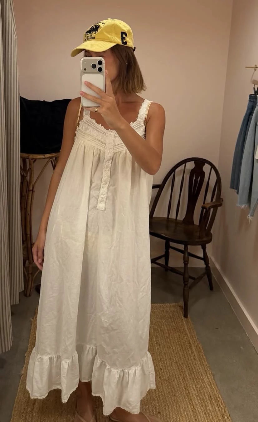 A woman takes a mirror selfie wearing a white sleeveless maxi dress with ruffled trim and a yellow baseball cap in a fitting room.