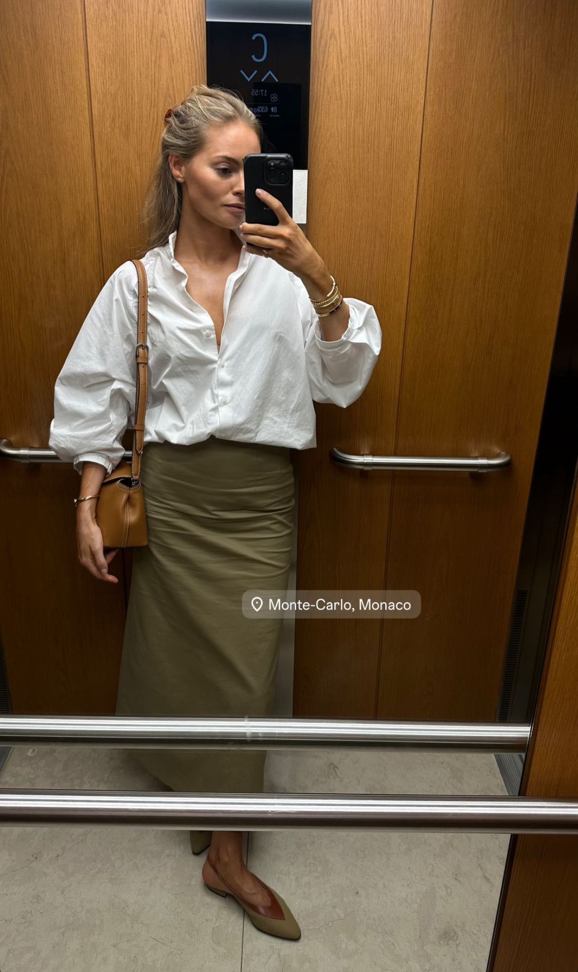 Woman taking a mirror selfie wearing a white shirt and green skirt in an elevator.