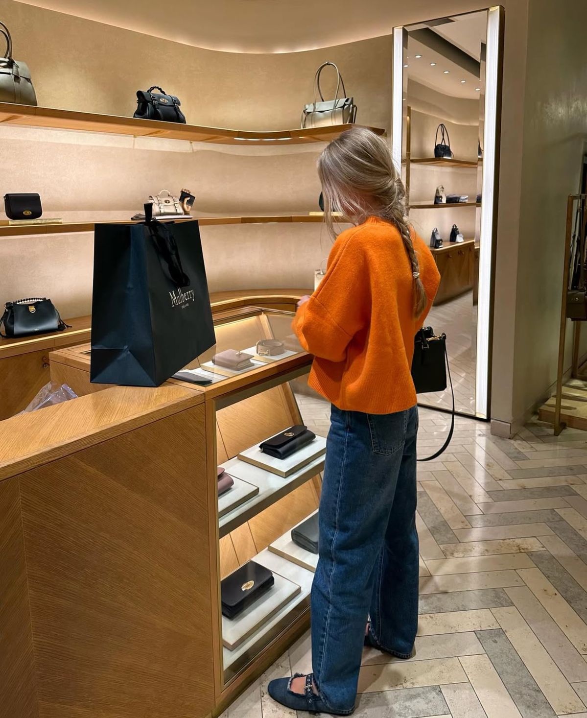 Person in an orange sweater and blue jeans standing in a store with shelves displaying handbags.