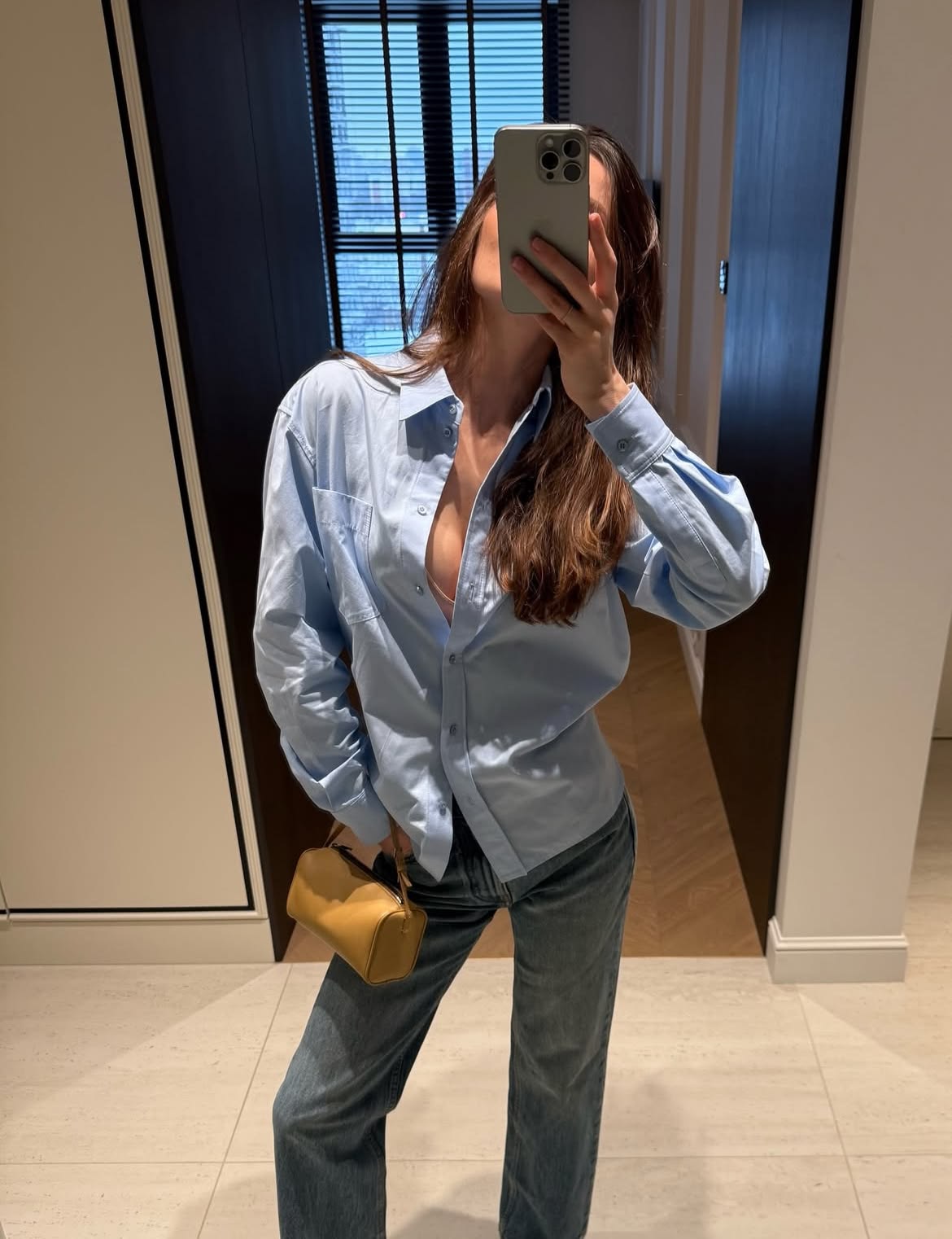 Person taking a mirror selfie wearing a light blue shirt and jeans, holding a yellow handbag.