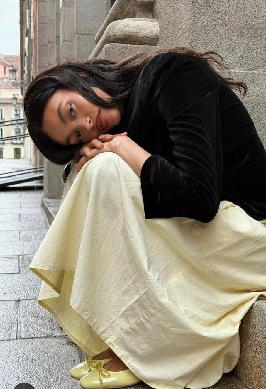 A woman sits slumped on stone steps outdoors, wearing a black velvet long-sleeve top and a flowing cream-colored skirt. She is wearing pale yellow shoes, and a black handbag rests beside her on the steps.