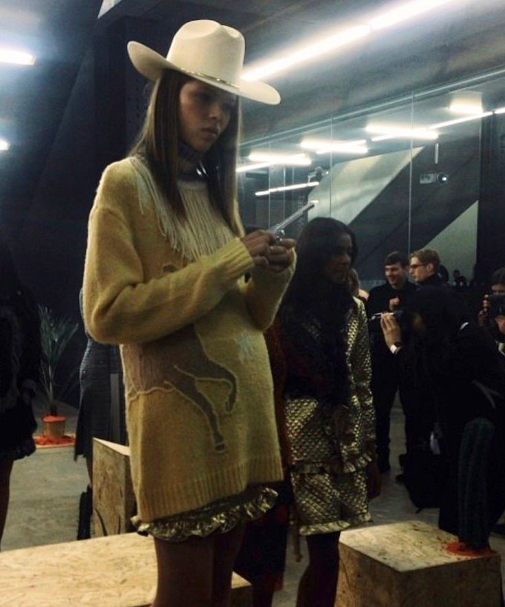 A model wearing a beige knit sweater dress with fringe and a graphic print, paired with a white cowboy hat, standing in a backstage fashion event setting.