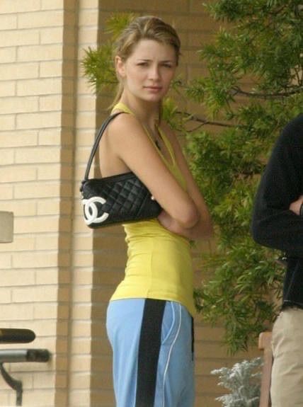 Woman in yellow top and blue pants holding a black handbag with a white logo, standing outdoors.