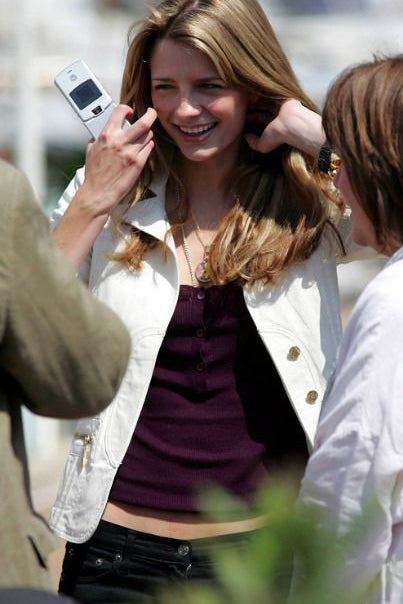 A woman outdoors wearing a white jacket over a dark purple top and black pants, holding a silver flip phone to her ear while another person adjusts her hair.