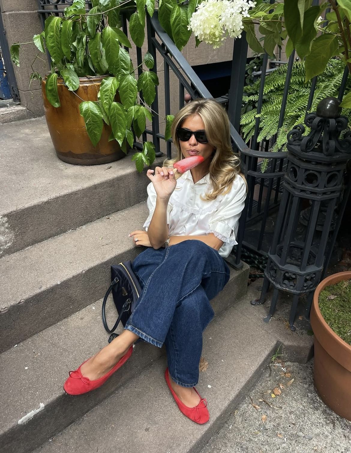 Woman sitting on steps eating a pink lollipop with plants and a black railing in the background.