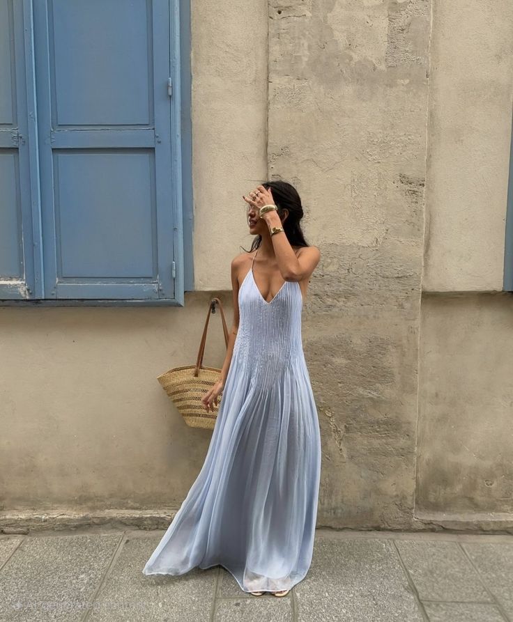 Woman in a light blue dress standing against a beige wall with blue shutters.
