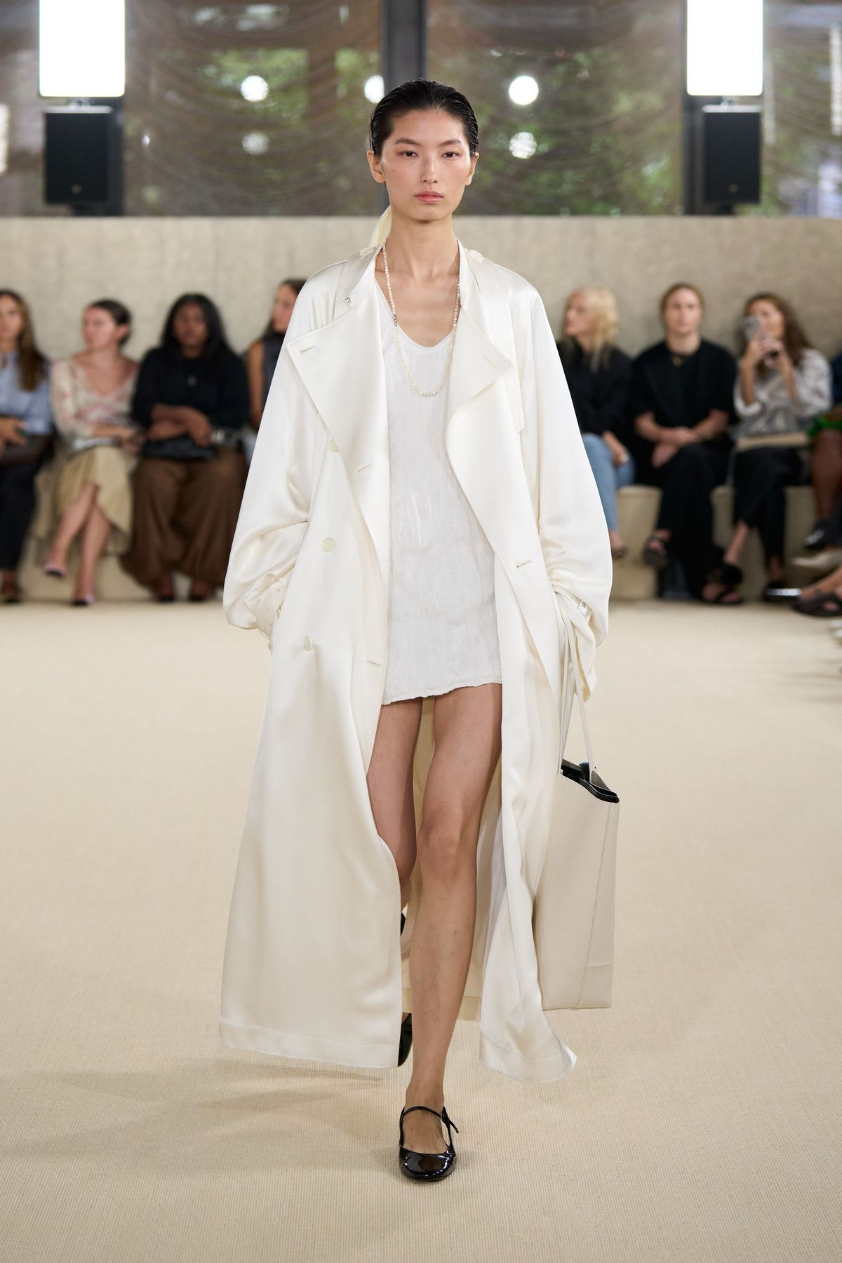 Model walking on a runway wearing a white outfit with a long white coat.