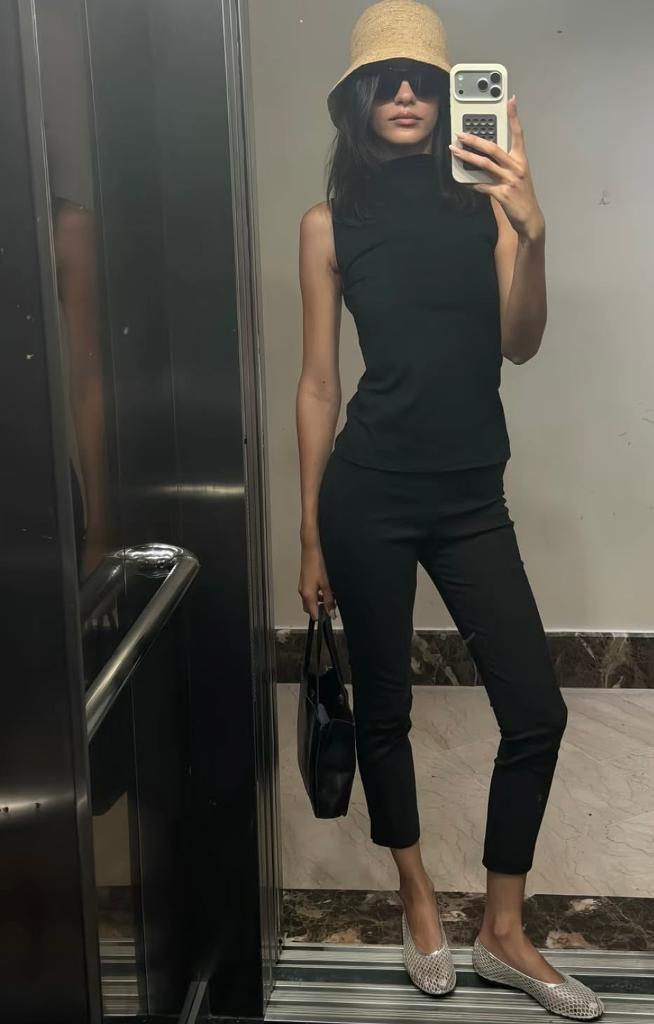 A woman takes a mirror selfie wearing a black sleeveless mock-neck top and black cropped pants, paired with a beige bucket hat, black handbag, and silver flat shoes.