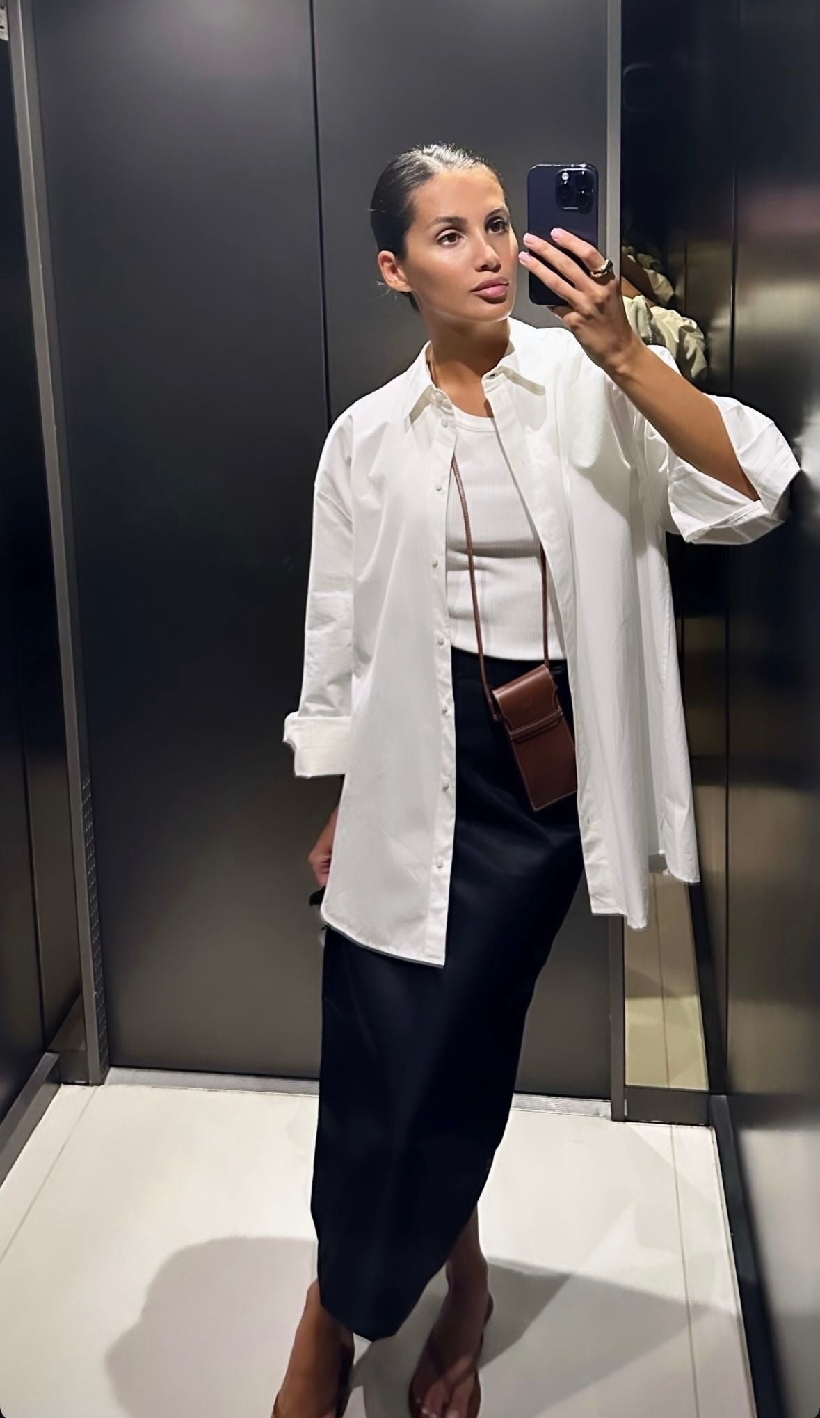 Woman taking a mirror selfie wearing a white shirt and dark skirt in a bathroom.