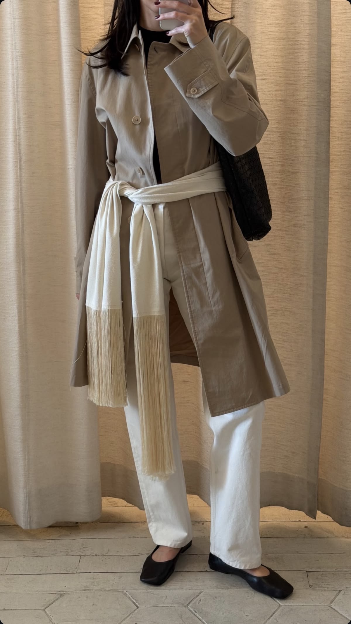 Person wearing a beige trench coat and white pants standing against a neutral curtain background.
