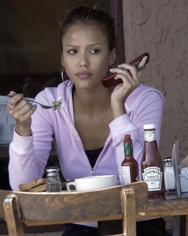 Woman in a pale pink zip hoodie holding a flip phone in a café.
