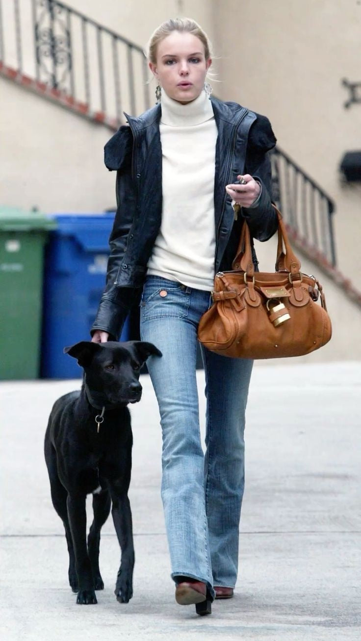 Woman in jeans and a leather jacket walking a black dog while carrying a tan bag.